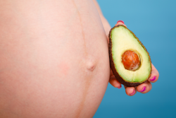 A person holding half an avocado with the seed intact next to a pregnant abdomen.