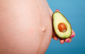 A person holding half an avocado with the seed intact next to a pregnant abdomen.