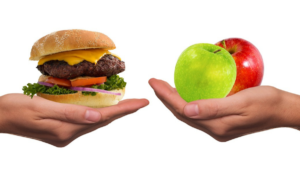 One hand holds a hamburger. A second hand holds two apples.